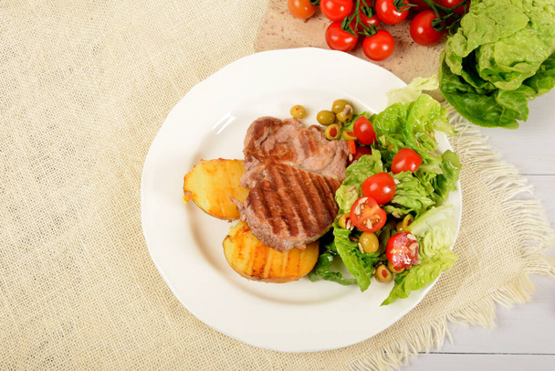 two pieces of grilled potato and grilled meat next to lettuce with tomatoes and olives on a white plate - Foto, Bild