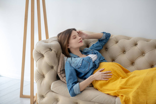 Young pregnant woman in a jeans jacket resting on the sofa - 写真・画像