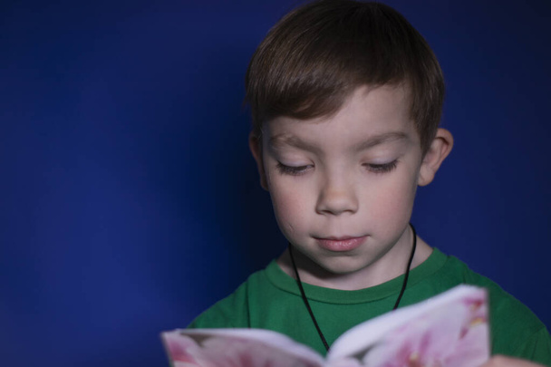 portret van een negen jaar oude blonde jongen het lezen van een boek in bloemen, op een blauwe achtergrond - Foto, afbeelding
