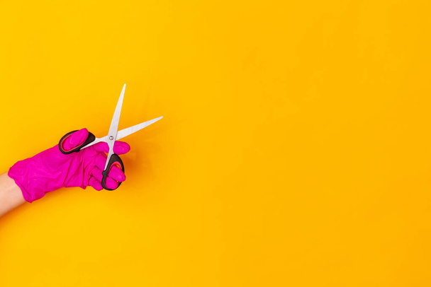 Hand in pink rubber glove holding sccissors isolated on yellow studio background with copyspace. - Фото, зображення