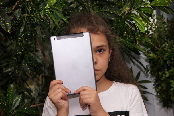 The girl holds a digital tablet in front of her face, covering half of her face. One eye. To spoil your eyesight. The concept of damage to vision from use of the gadgets . - Foto, afbeelding