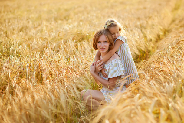 Μια όμορφη κοκκινομάλλα νεαρή μητέρα και η κόρη της αγκαλιάζονται σε ένα χωράφι με σιτάρι μια ηλιόλουστη μέρα.. - Φωτογραφία, εικόνα