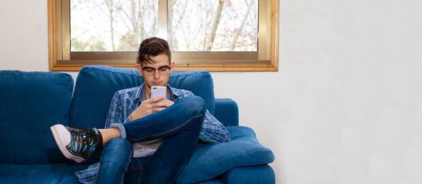 jeune homme ou adolescent avec téléphone portable assis sur le canapé à la maison ou à l'appartement - Photo, image