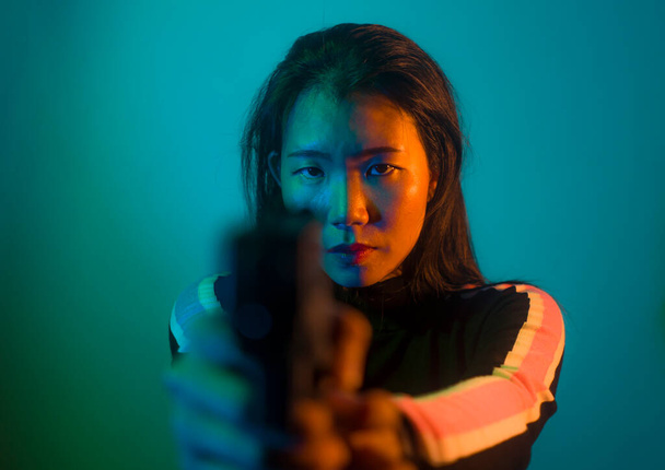 edgy cinematic portrait of young attractive and dangerous special agent woman or Asian mobster girl holding handgun pointing the gun fierce in Hollywood movie style - Foto, Imagem
