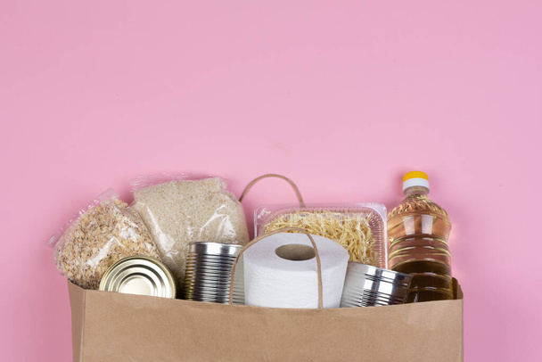 Paper bag with crisis food supply for the period of quarantine isolation on pink background with copy space, rice, pasta, oatmeal, canned food, toilet paper. Food delivery, donation during coronavirus - Foto, Imagen
