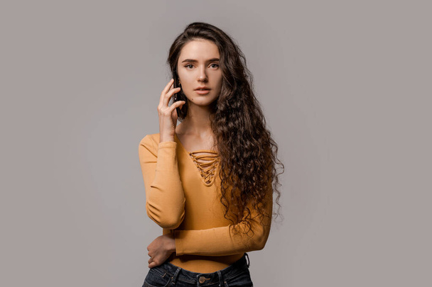 Chatting on mobile cell phone, online internet technology concept. 5g connecting and communication. Curly cute concentrated woman have a call dressed in yellow over grey background - 写真・画像