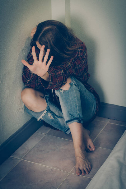 Μια γυναίκα με φόβο κάθεται στο πάτωμα στον τοίχο και καλύπτει το πρόσωπό της. - Φωτογραφία, εικόνα