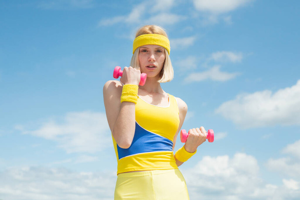 Healthy young attractive sportswoman wearing yellow with blue training uniform demonstrating exercises with pink dumbbells on sky background with clouds. Fashion and Trends in Sport. Health and - Foto, Bild