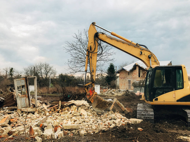 Bulldozer clearing land from old bricks and concrete from walls with dirt and trash. Backhoe machinery ruining house. Excavator destroying brick house on land in countryside. - Photo, Image