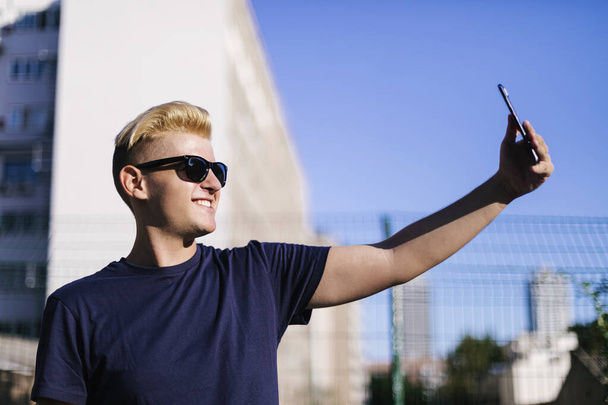 Молодий чоловік в сонцезахисних окулярах приймає селфі на відкритому повітрі
 - Фото, зображення