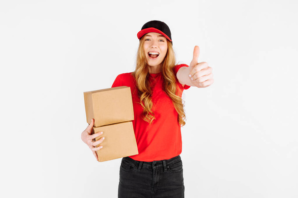 Happy courier girl in uniform holding cardboard boxes and showing a thumbs up gesture on a white background. The concept of home delivery - Фото, изображение