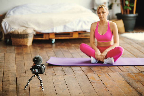 young fit blogger girl seating on  yoga  mat in sport outfit with laptop doing online  workout at home, recording online aerobic tutorials from home, exercise on camera - Photo, Image