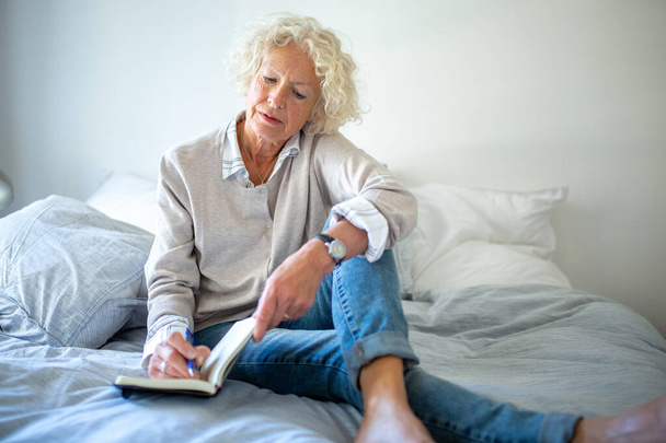 Портрет літня жінка сидить на ліжку пише в щоденнику
 - Фото, зображення