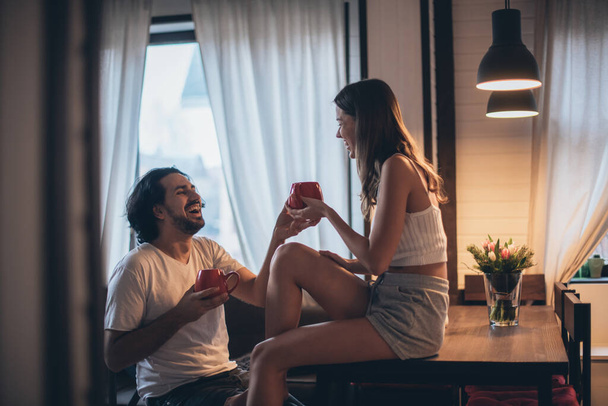 Loving couple in the evening in the living room at home. A young family is drinking tea, a girl is sitting on a table. Romantic family evening at home, dim light. - Photo, Image