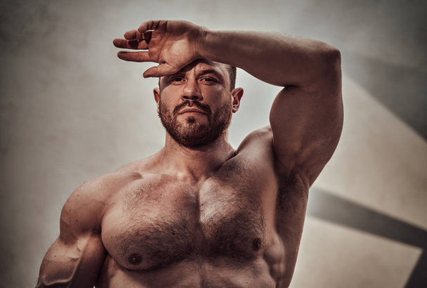 Zmotywowany silny mężczyzna pozujący w ciemnym studio, gotowy do rozpoczęcia treningu i pokazania swoich wielkich bicepsów zakrywając oczy przed światłem słonecznym - Zdjęcie, obraz