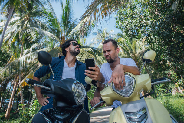 Два человека на мотоциклах ищут дорогу на карте в телефоне на тропическом острове. Ребята туристы смотрят на штурмана на смартфоне на велосипедах на дороге в пальмовой роще
 - Фото, изображение