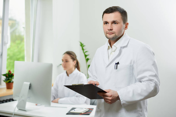 Egy komoly sebész fehér köpenyben tart egy fekete írótáblát, ahol dokumentumokat elemez a kórházban. Egy női orvos ül az asztali számítógép előtt a szoba sarkában..  - Fotó, kép