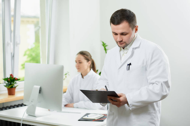 Een serieuze chirurg met borstelharen in een witte jas houdt een zwart klembord vast en vult de patiëntenkaart in een ziekenhuis. Een vrouwelijke arts zittend achter de desktop computer in de hoek van de kamer  - Foto, afbeelding