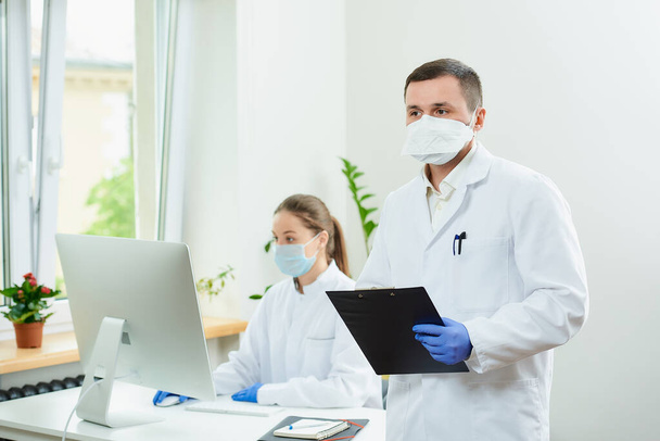 A serious surgeon wears a respirator and disposable medical gloves holding a black clipboard with documents in a hospital. A female doctor sitting in front of the desktop computer in the corner.   - Photo, Image