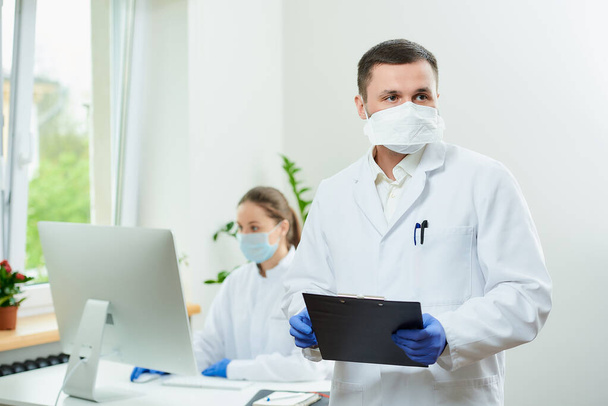 Un cirujano con cerdas usa un respirador sostiene un portapapeles negro y espera en un hospital. Una doctora sentada frente a la computadora de escritorio en la esquina de la habitación.  - Foto, imagen