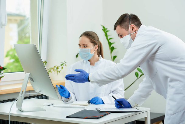 Ένας χειρουργός και ένας ακτινολόγος με ιατρικές μάσκες προσώπου συζητούν ακτινογραφίες ενός ασθενή στην οθόνη του υπολογιστή. Μια γυναίκα γιατρός κάθεται μπροστά στον επιτραπέζιο υπολογιστή, ένας θεραπευτής μένει κοντά. - Φωτογραφία, εικόνα