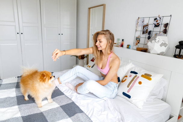A happy blond woman is sitting on the bed with her dog of breed Spitz. Portrait of a woman and a dog. - Foto, Imagem
