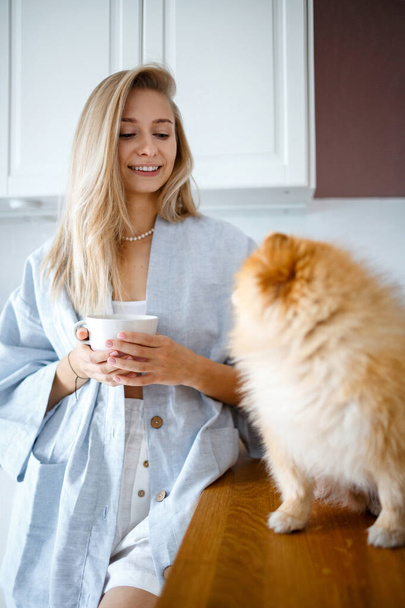Happy blonde woman is drinking tea in the kitchen, next to her is her Pomeranian breed dog. The friendship of people and animals. - 写真・画像