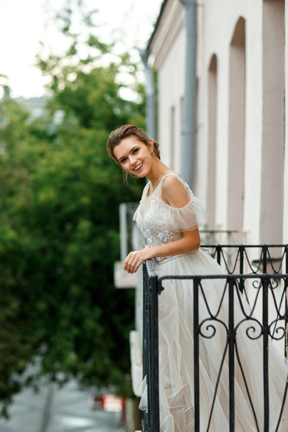 Krásná mladá nevěsta ve svatebních šatech stojí na balkóně a usmívá se. - Fotografie, Obrázek