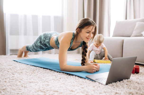 Sport at home. Young attractive woman doing sports at home and watching a video fitness lesson on a laptop, and her little baby girl - Fotoğraf, Görsel