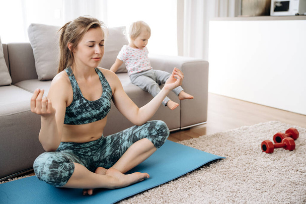 Sport en yoga thuis. Moeder oefent yoga zitten in een lotus positie op de vloer, en haar kleine schattige dochter speelt in de buurt - Foto, afbeelding