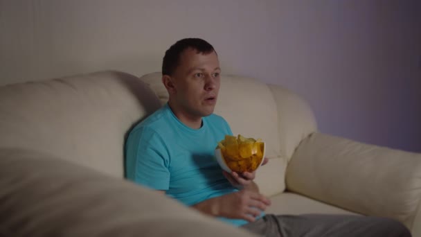 The man is shocked at the transmission he saw on TV, sitting on the sofa with a bowl of chips - Imágenes, Vídeo