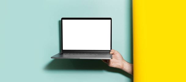 Close-up of male hand holding laptop with mockup between two studio backgrounds of yellow and aqua menthe colors. - Фото, изображение