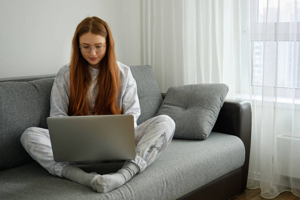 Red-haired girl in pajamas and glasses sits on a sofa and types on a laptop - Foto, Bild