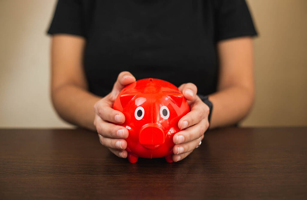 a latin girl sit with sit in front of a desk put in bills in a red Piggy bank - Photo, Image
