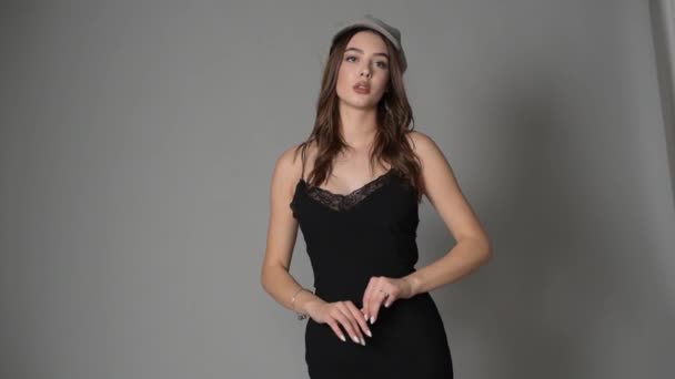 Glamorous young woman wears fashionable dress posing with fluttering hair at grey wall. Girl put forward hand in a black dress and a gray cap. Beautiful young girl with brown hairs. portrait - Footage, Video