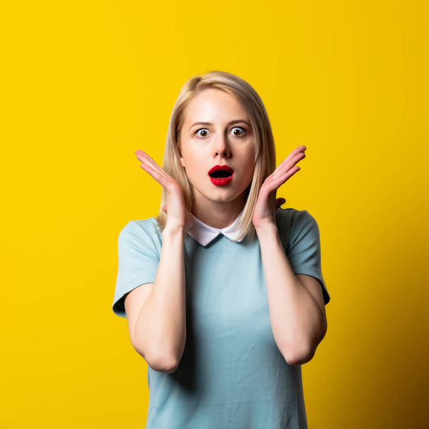 Surprised blonde girl in blue dress on yellow background - Photo, Image