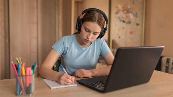 Teenager Mädchen zu Hause mit Kopfhörern schreibt mit einem Stift in einem Notizbuch, hört Lektionen und lernt im Internet, ein Laptop mit dem Internet. Fernstudium während der Quarantäne, zu Hause bleiben. - Foto, Bild