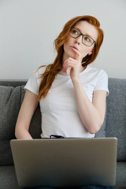Redhead girl sits on a sofa in a white T-shirt and glasses, with a laptop, looks thoughtfully at the top, putting finger to lips - Fotó, kép