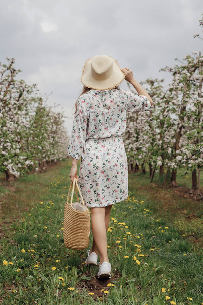 Девушка в соломенной шляпе и платье в саду. Женщина с сумкой гуляет по саду между деревьями с цветами. Яблочный сад весной. Фотосессия в природе
. - Фото, изображение