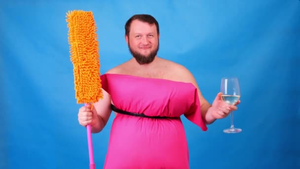 Funny bearded guy in a pink pillow dress with a mop and a glass of wine on a blue background. Crazy quarantine. Funny house cleaning. Fashion 2020. Put on a pillow. Challenge 2020 due to house - Footage, Video