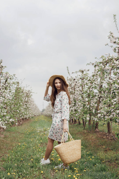 Tyttö olkihatussa ja mekko puutarhassa. Nainen pussin kanssa kävelee puutarhassa puiden välissä kukkia. Omenatarha keväällä. Photo ampua luonnossa. - Valokuva, kuva