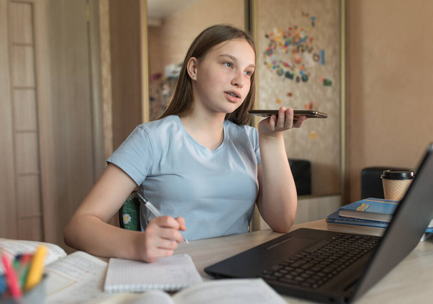 Teenage girl recording audio call on phone, at home homework Internet laptop, social connections 4G 5G. Distance education training quarantine. Background books lectures on school institute. - Zdjęcie, obraz