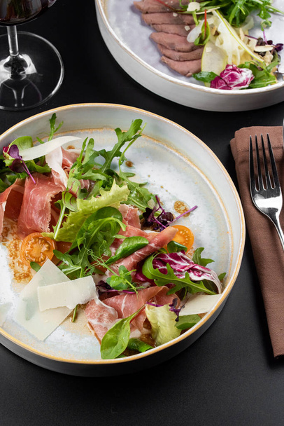 tasty fresh dishes with green salad and meat prosciutto slices on white plate on table background - Photo, Image