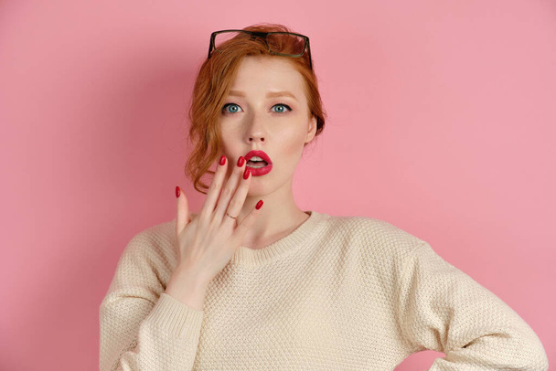 A beautiful red-haired girl with red lipstick and blue eyes stands on a pink background and looks in surprise at the frame, bringing hand to lips - Zdjęcie, obraz