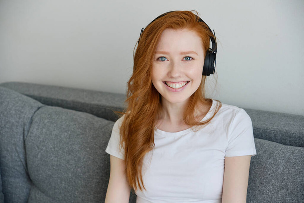 The red-haired girl in a white T-shirt and headphones sits on the sofa and smiles openly into the frame. - Foto, Bild