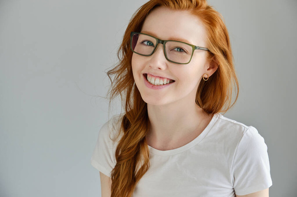 Red-haired girl in a white T-shirt and glasses stands on a gray background in half-turn and smiles - Foto, Imagem