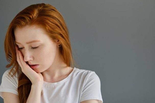 A red-haired girl in a white T-shirt is standing in a half-turn on a gray background with her head down and palm resting on her face - Foto, Imagen