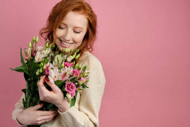 Cute red-haired girl stands on a pink background, with a smile presses a bouquet to herself, looking at him - Fotó, kép