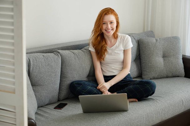Een roodharig meisje zit gekruiste benen op een bank in huis kleren met een laptop en glimlacht breed - Foto, afbeelding