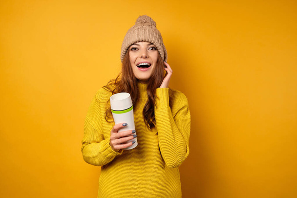 A brunette in a yellow sweater stands on a yellow background with a thermos in hands and looks joyfully into the frame, mouth open - Foto, immagini